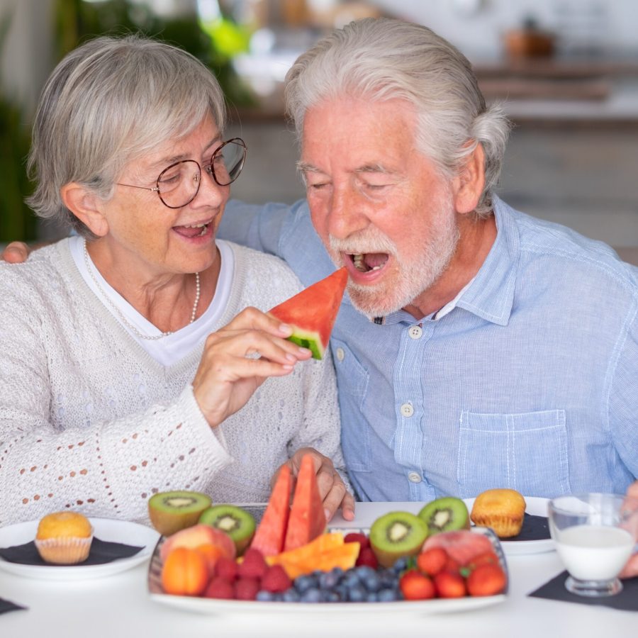 Cheerful senior couple at home having breakfast together with muffin, milk and fresh seasonal fruit, healthy eating concept
