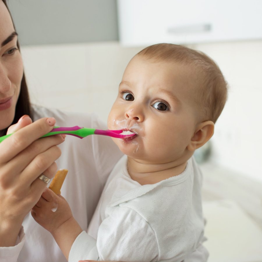 Close up of lovely newborn boy with big brown eyes eating porridge, looking in camera while mother feeding him with spoon and holding kid on hands