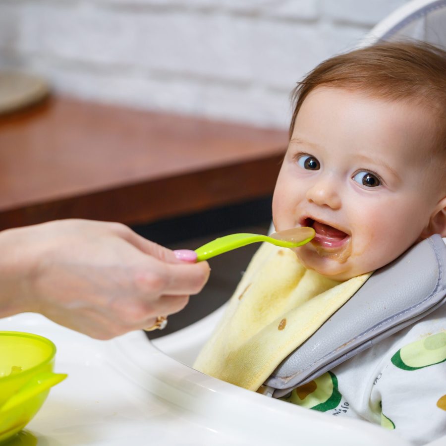 A nine month old smiling baby sits at a white table in a highchair and eats with a spoon from a bowl. Mom feeds the baby from a spoon. Blurred background. Healthy food for children. Children food.
